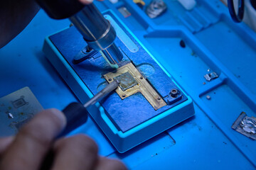 The hands of cellphone technology device maintenance engineer work on the smartphone microprocessor pins with flux, hot air heater and soldering iron. Close-up macro photo, selective focus
