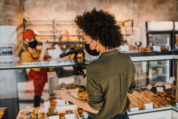 Beautiful African American woman with protective face mask buying fresh bread in bakery.