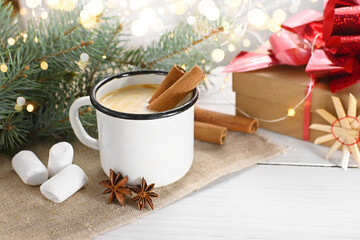 Eggnog with cinnamon, anise and nutmeg in a white mug on a wooden table near the branches of a Christmas tree and decorative toys.