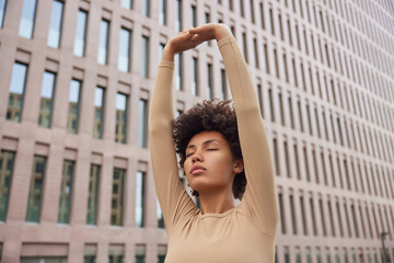 Athletic calm woman with curly hair raises arms over head does stretching exercises wears beige...