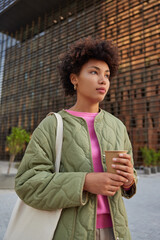 Young dreaming curly haired woman holds disposable cup of tasty coffee enjoys good weather while strolling in city carries bag focused forward spends leisure time outdoors walks in downtown.