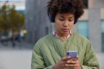 Cropped image of hipster girl listens audio record with high volume enjoys music on radio via headphones concentrated at smartphone screen poses outside against blurred background. Leisure time