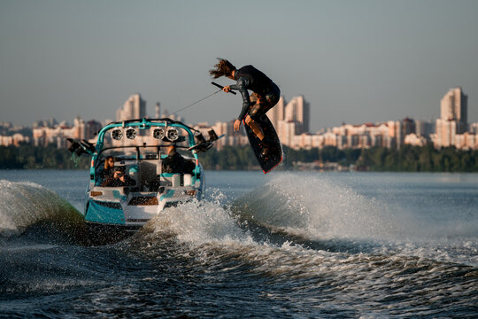energy wakeboarder moving fast behind boat holding rope and masterfully jumping high on splashing river wave.