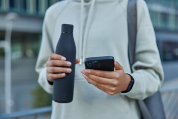 Cropped shot of sportswoman in casual hoodie holds bottle of water and mobile phone checks fitness activity carries rolled mat poses against blurred background takes break during cardio workout