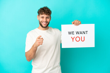 Young handsome caucasian man isolated on blue background holding We Want You board and pointing to...