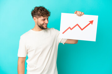 Young handsome caucasian man isolated on blue background holding a sign with a growing statistics arrow symbol with happy expression