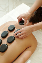 Hands of masseuse putting hot spa stones on female back during healthcare and beauty procedure in luxurious salon