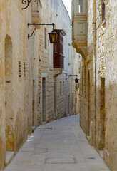 Typical Mdina street. Narrow medieval street of Mdina, also known as "Silent city", paved with stone slabs, surrounded with yellow limestone walls with vintage lanterns and enclosed balcony.