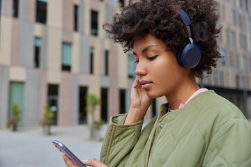 Sideways shot of pretty woman listens audio book via headphones installs media app on cellular...