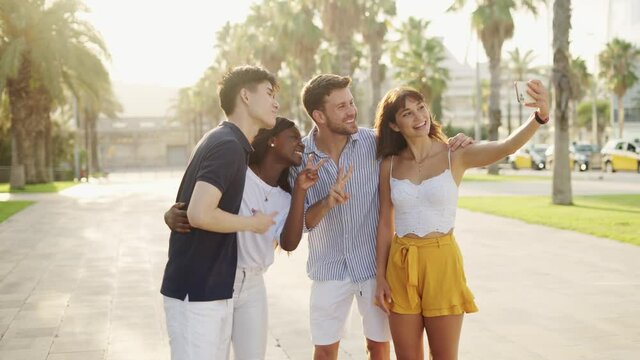 Positive young multi-ethnic friends making selfie on smartphone while standing on sunny embankment