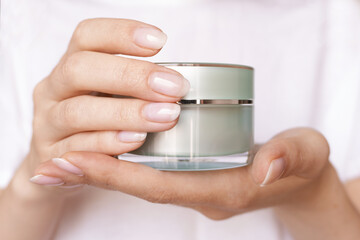 Female hands with natural pink manicure holding a transparent cream jar. Woman in white holds beauty product. Concept of body care and skin protection. Moisturizing lotion closeup. Facial skincare