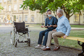 Talkative caring young nurse wearing face shield chatting with aged man, recovering patient while sitting together on the bench in park near hospital