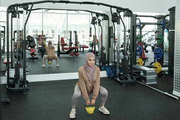 Young strong Muslim woman with heavy weight exercising on knee bent legs in large fitness center
