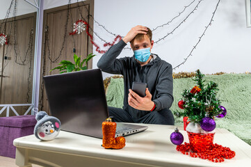 Angry men in grey hoodies and medical mask at home and talking on the phone and clutching their heads during Christmas, trouble