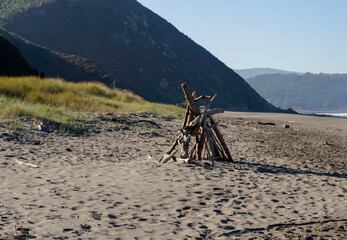 Conjunto de maderas y palos formando una cabaña y una hoguera en medio de la arena de la playa.