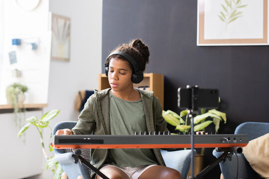 Cute preteen biracial schoolgirl in headphones playing piano keyboard while sitting in domestic room