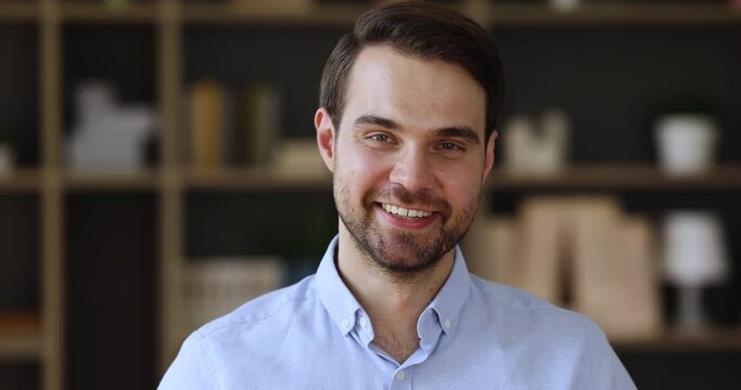 Head shot portrait of young adult handsome bearded businessman posing indoors show white teeth in sincere smile. Positive millennial male office employee manager look at camera feel happy motivated