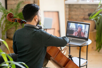 Back view of young music teacher playing cello in front of laptop with cute little schoolboy on screen © pressmaster