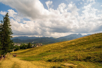 Wide angle view of green meadow countryside field with A shaped house and people hiking on a trail...