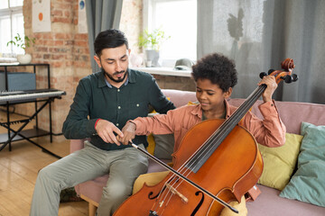 Cute biracial boy playing cello while sitting on couch next to his teacher during lesson of music in home environment - Powered by Adobe