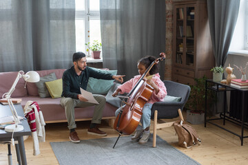Young Caucasian man with paper pointing at biracial schoolgirl with cello during home lesson of classical music