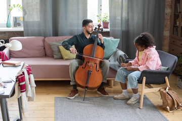 Young teacher of music showing how to play cello to biracial pupil sitting in armchair in front of him © pressmaster