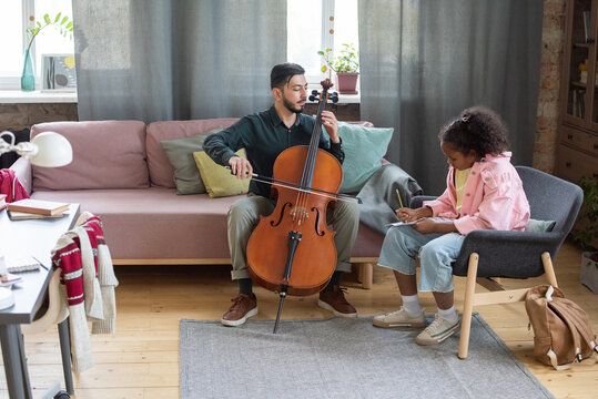 Young teacher of music showing how to play cello to biracial pupil sitting in armchair in front of him