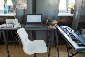 White chair of student standing between table with laptop and piano keyboard in home environment