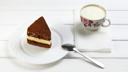 Triangular piece of brown cake on plate, spoon and cup with kefir as breakfast on wooden white surface
