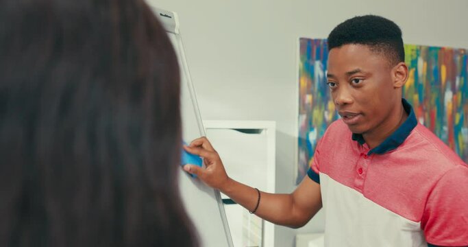 Dark-skinned teacher holds tablet in hands stands at blackboard with young handsome student, boy holds marker in hand, looks at graph drawn by translator pointing, wipes the board school classroom