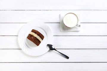 Piece of brown cake on plate, spoon and cup with kefir as breakfast on wooden white surface in flat lay