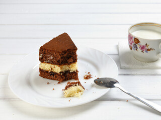 Piece of cake on plate in process of eating part, spoon and cup with milk product on white wooden table, close-up
