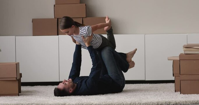 Happy young father lying on floor carpet, lifting in air small adorable child daughter, having fun playing together in modern living room, celebrating moving into new apartment dwelling, real estate.