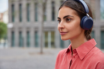 Sideways shot of pretty young woman listens music via headphones looks pensively forward dressed in pink shirt strolls in megapolis enjoys audio playlist stands outdoors against blurred background