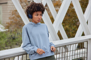 Pensive young woman with curly hair poses at bridge wears blue sweatshirt and trousers listens motivational music via wireless headphones focused into distance. People leisure and hobby concept