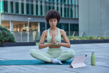 Young relaxed woman does yoga at mat sits in lotus pose keeps palms pressed together eyes closed breathes deeply watches tutorial video on modern tablet poses at mat outdoors inn urban place