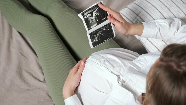 Pregnant woman in white shirt and tights looks at ultrasound picture holding in hand and strokes belly sitting on bed close upper view