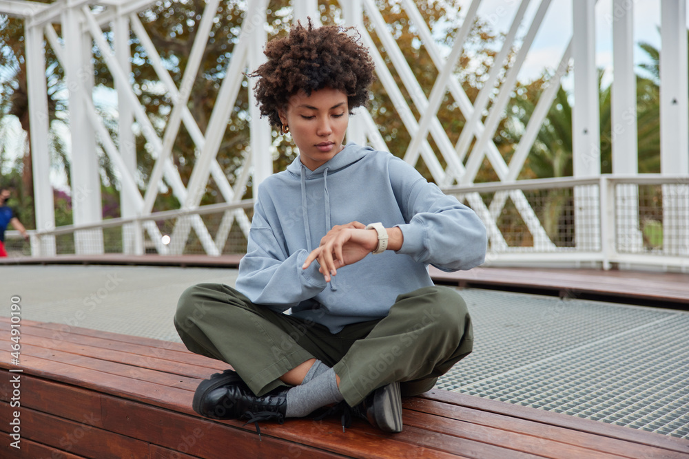 Wall mural Outdoor shot of teenage girl with curly bushy hair sits crossed legs wears casual hoodie trousers and shoes looks at wrist watch checks time poses at bridge during daytime waits for someone. - Wall murals