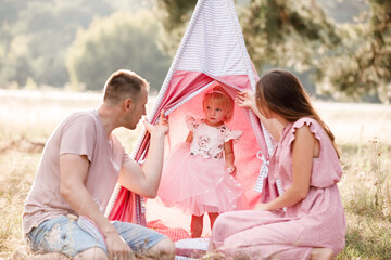 Mom, dad and little daughter are sitting next to wigwam decoration in the park. Family spending time outdoor in summer, having fun together. Girl are dressed in pink dress. © Andriy Medvediuk