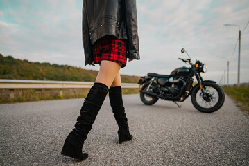 motorcyclist woman in mini and leather jacket walking to retro-styled motorcycle. Attractive female driver in jackboots alone on roadway. Trip, cafe racers, speed, freedom concept.