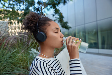 Sideways shot of curly haired thirsty sporty woman drinks refreshing water from bottle tired after workout listens music via headphones wears striped jumper poses outdoors rests after jogging