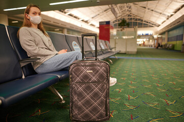 A young woman wearing a medical mask sits in the waiting room at the train station. She has a suitcase next to her. There's no one around. Travel during quarantine, social distancing, global pandemic.