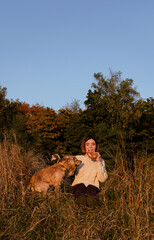 Young woman with short hair sitting on dry grass hill during sunset with her English cocker spaniel dog, eating pastry.