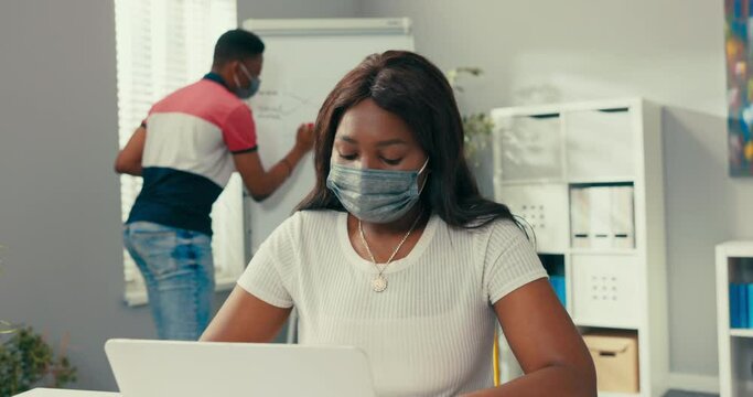 A busy man stands at the blackboard holding a tablet, looking for the right graphic, preparing a diagram, a beautiful woman sits at her desk entering data into the system needed to create the diagram