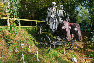 spooky halloween decoration with two real size skeletons riding in a coach with horses in the woods, skeletons sticking out of the ground - 464920531