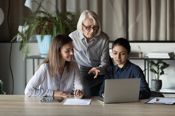 Group of two multiethnic females students sit at desk on workshop seminar listen to experienced mature woman teacher. Aged lady team leader boss explaining work to younger staff members subordinates