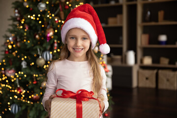 Portrait of happy beautiful small cute child girl in red hat holding wrapped gift box with...