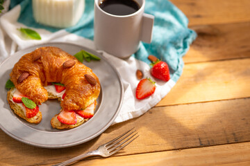 Delicious nutritious breakfast for one person - a cup of fragrant coffee, milk, fresh croissant with butter, strawberries, green mint leaves, almonds. Wooden texture.