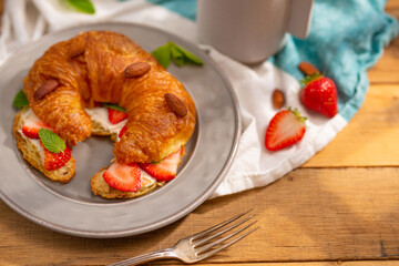 One delicious croissant with butter, strawberries, almonds, mint leaves, a cup of coffee and milk. Sunlight. Beautiful composition. Romance, festive mood, sweet food.