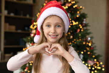 Head shot portrait of happy cute little 7s child girl in red santa hat showing heart sign with...